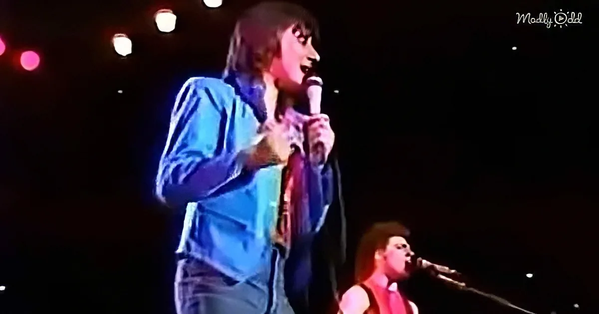 A person in a blue jacket holds a microphone on stage, illuminated by stage lights.