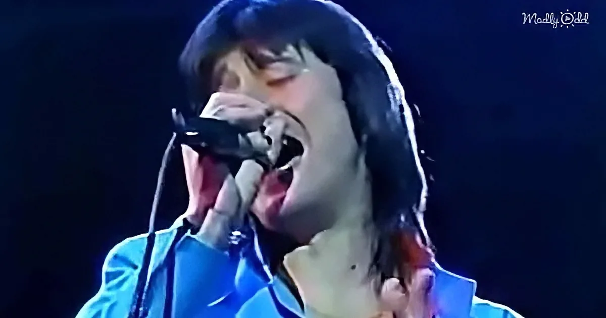 A person in a blue shirt sings passionately into a microphone against a dark background.