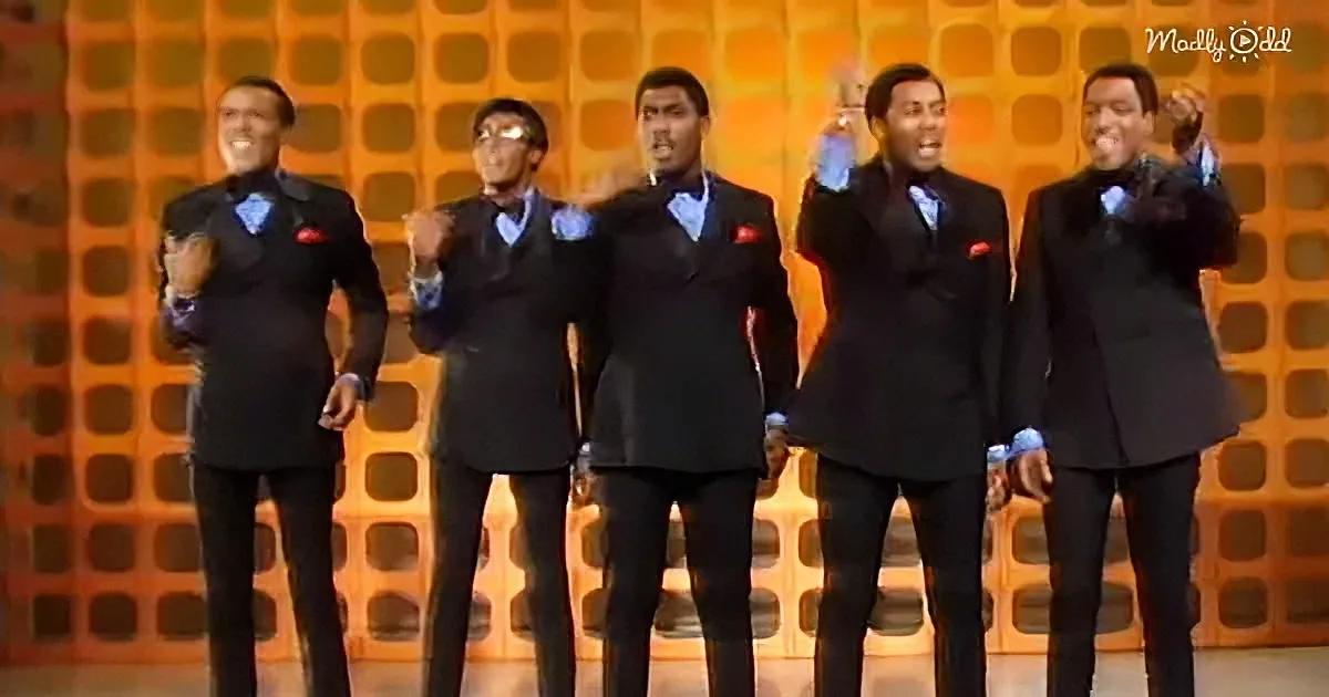 A group of five men in matching black suits and bow ties perform on stage with a vibrant orange background.
