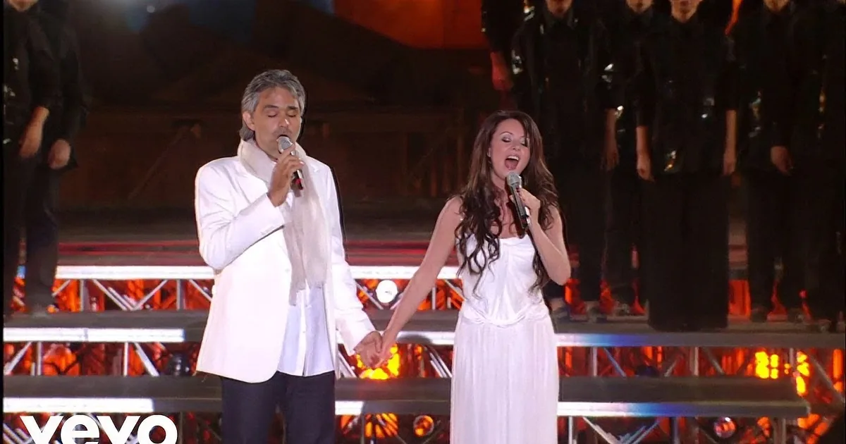 A man and woman in white sing on stage, holding hands, with a band and choir behind them.