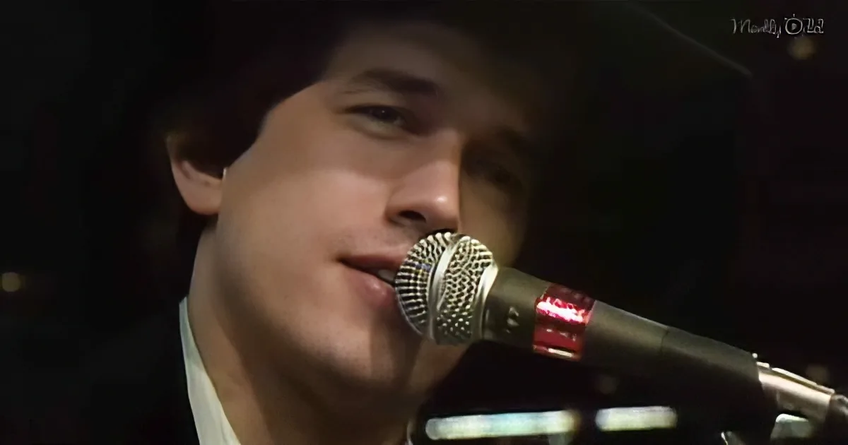 A person singing into a microphone, wearing a white shirt, with a dark background.