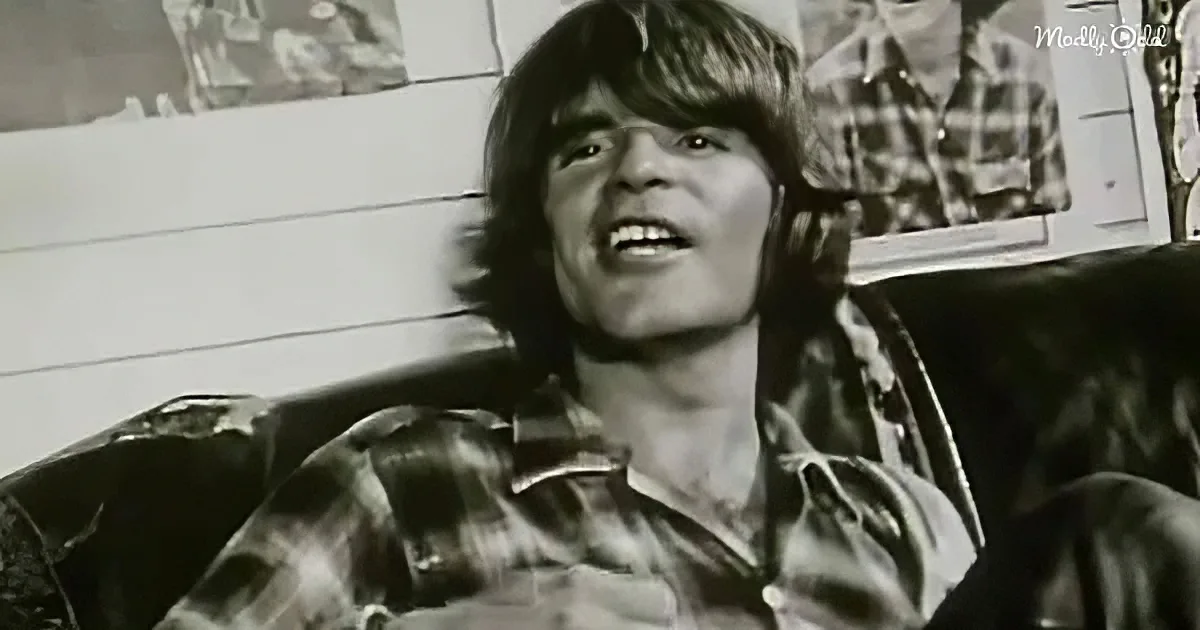 A person with long hair sits on a couch, wearing a patterned shirt, in a room with posters on the wall.