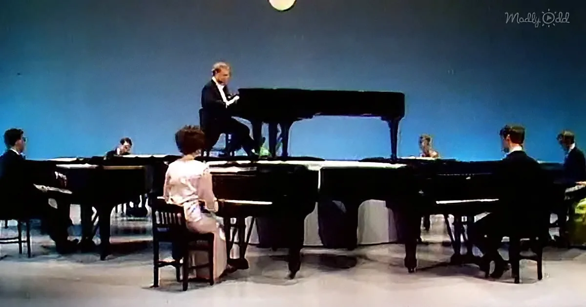 A man plays a grand piano on stage under a full moon, surrounded by seated musicians.