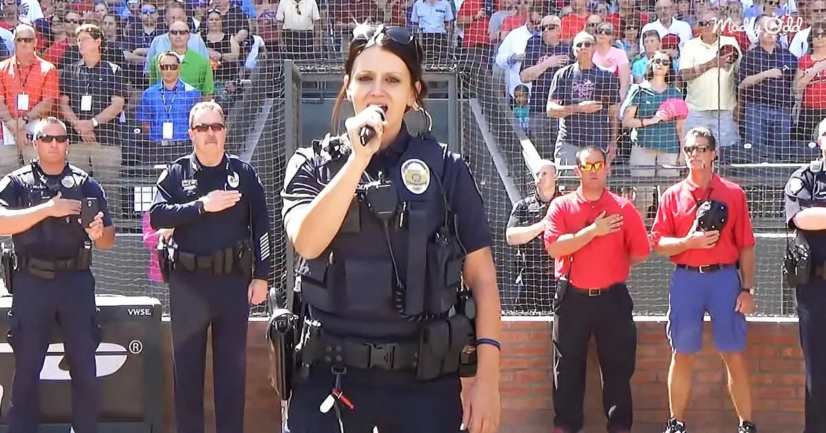 A woman in a police uniform sings into a microphone while others stand with hands over hearts.