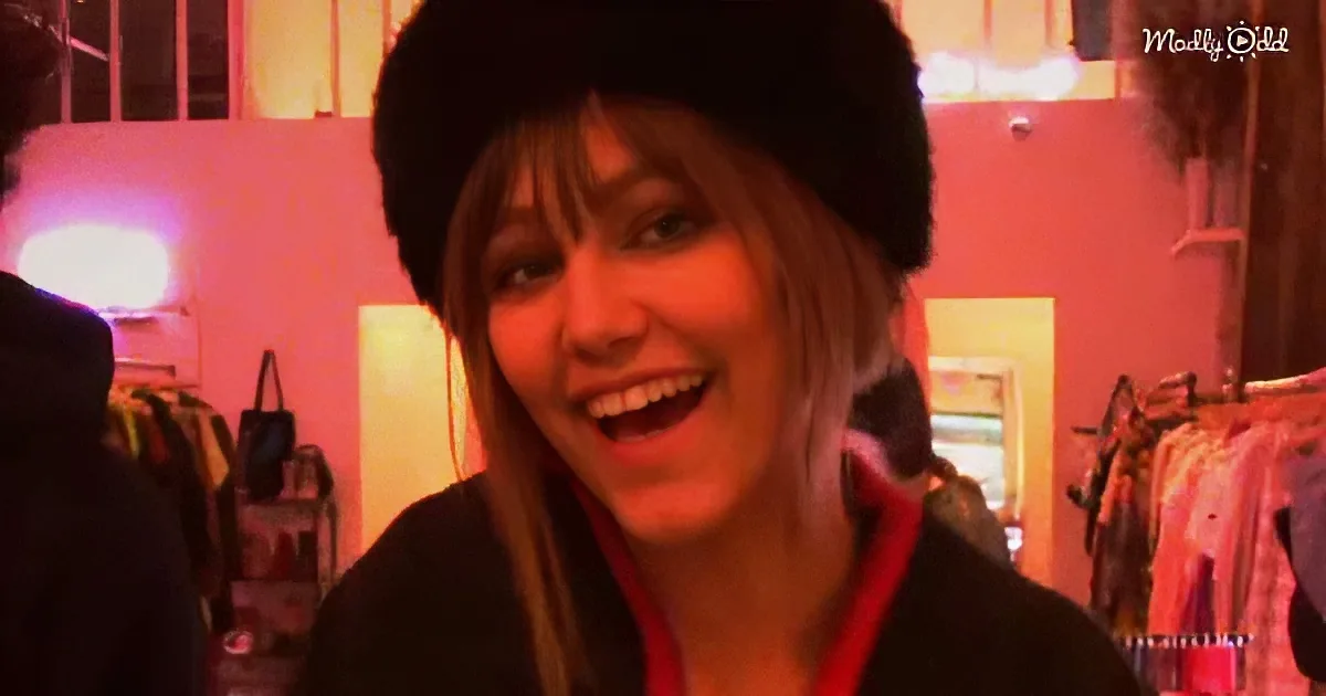 A smiling woman with a fur hat, red scarf, and dark jacket in a warmly lit room with clothing racks.