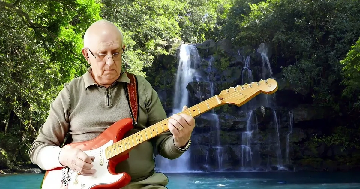 An older man plays a red electric guitar in front of a waterfall surrounded by lush greenery.
