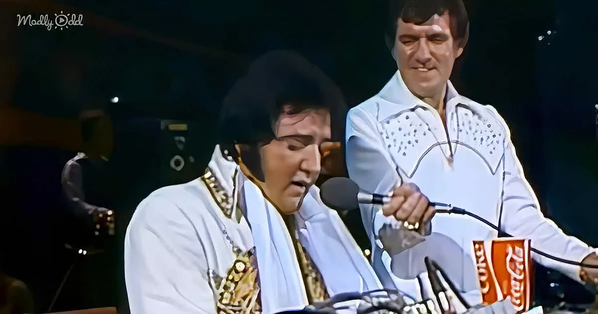 Elvis Presley in white stage outfit sings into microphone, another man in similar attire stands nearby.