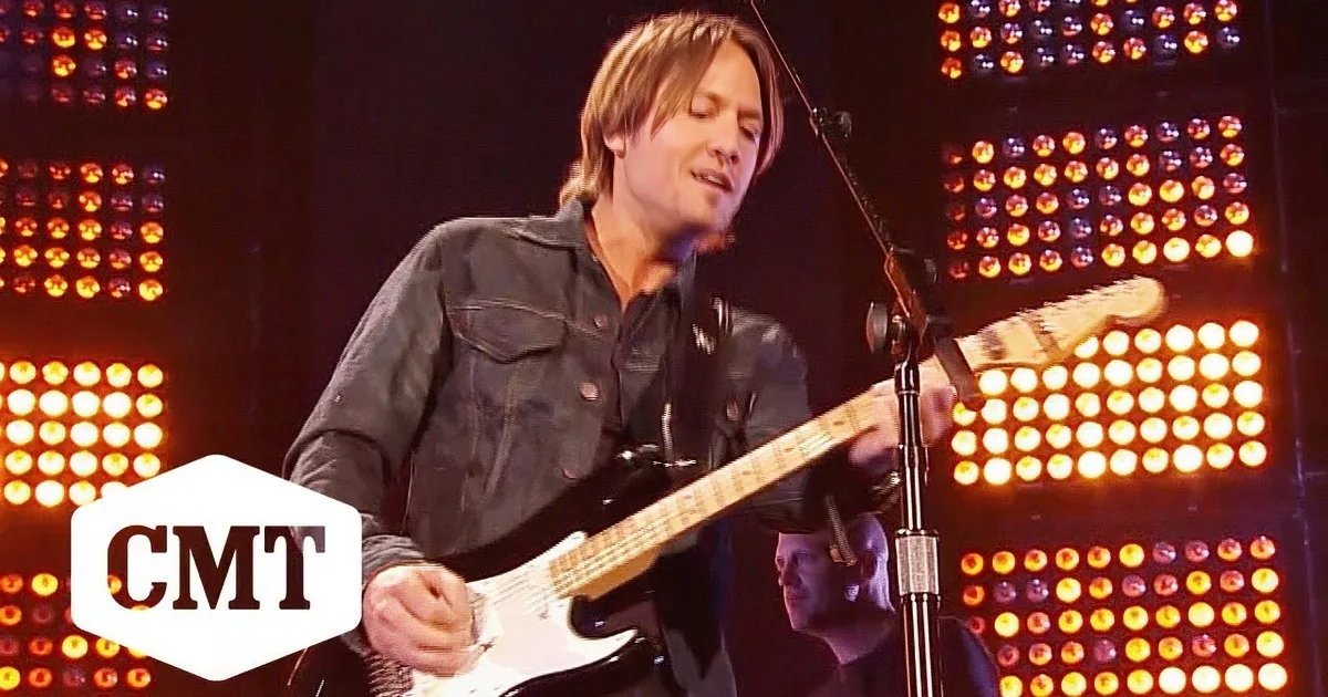 A man plays a white electric guitar on stage with bright orange lights behind him.