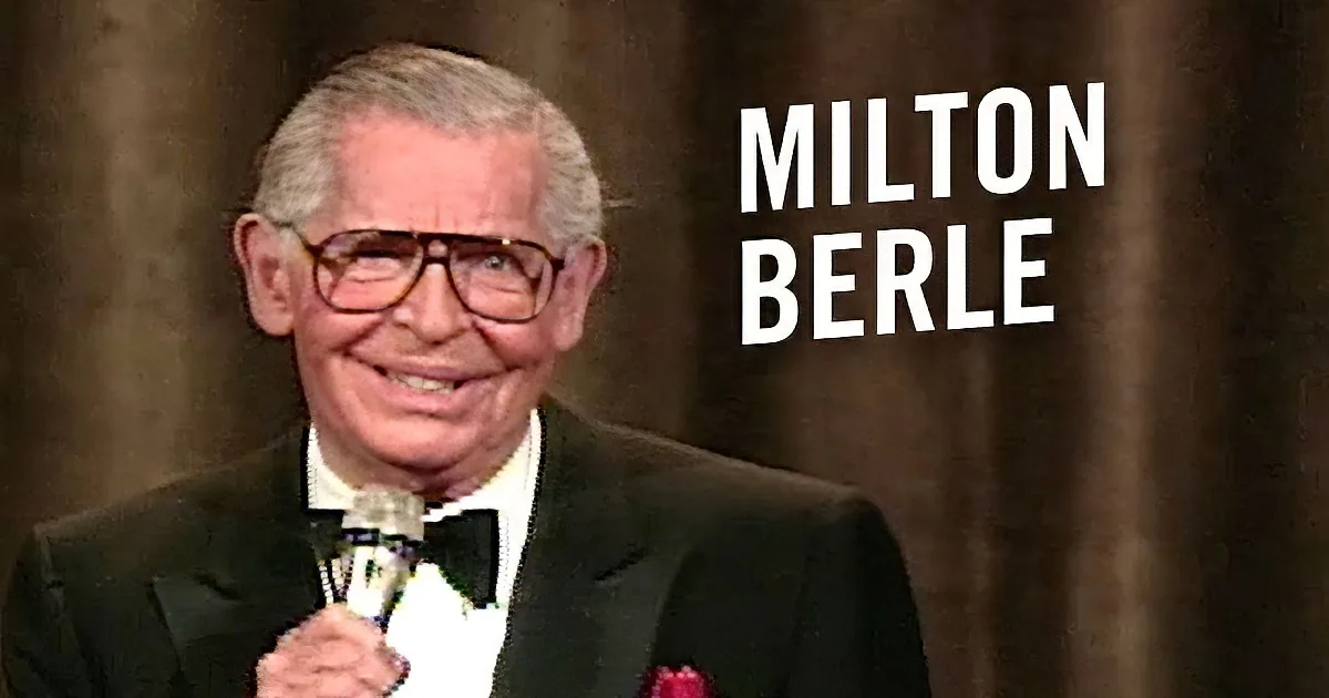An older man in a tuxedo smiles while holding a microphone on stage.