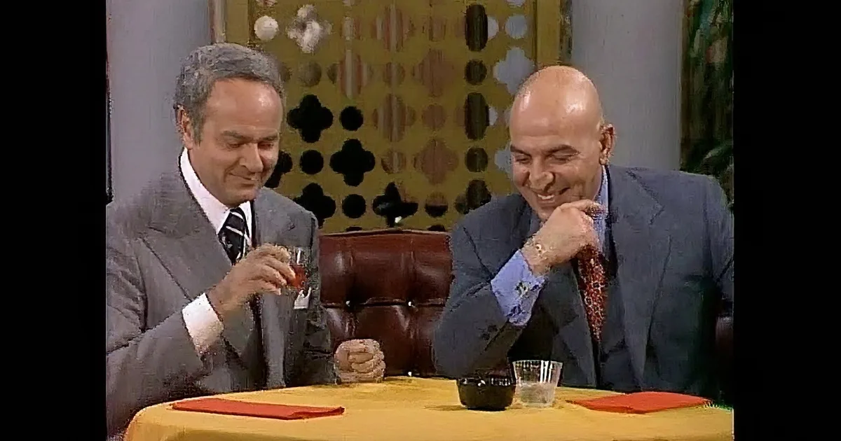 Two men in suits, one with a gray suit and the other in blue, are smiling and appear to be in a conversation.