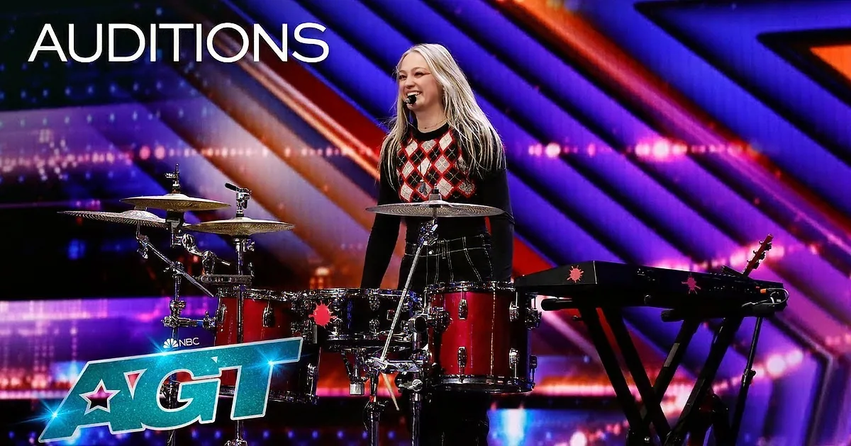 A young woman plays drums on a stage with a colorful background.