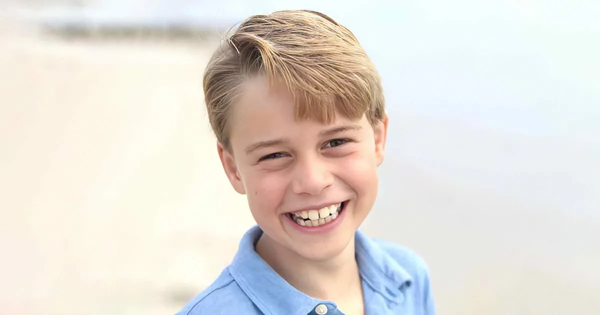 A young boy with blonde hair, wearing a blue shirt, smiles brightly against a blurred beach background.