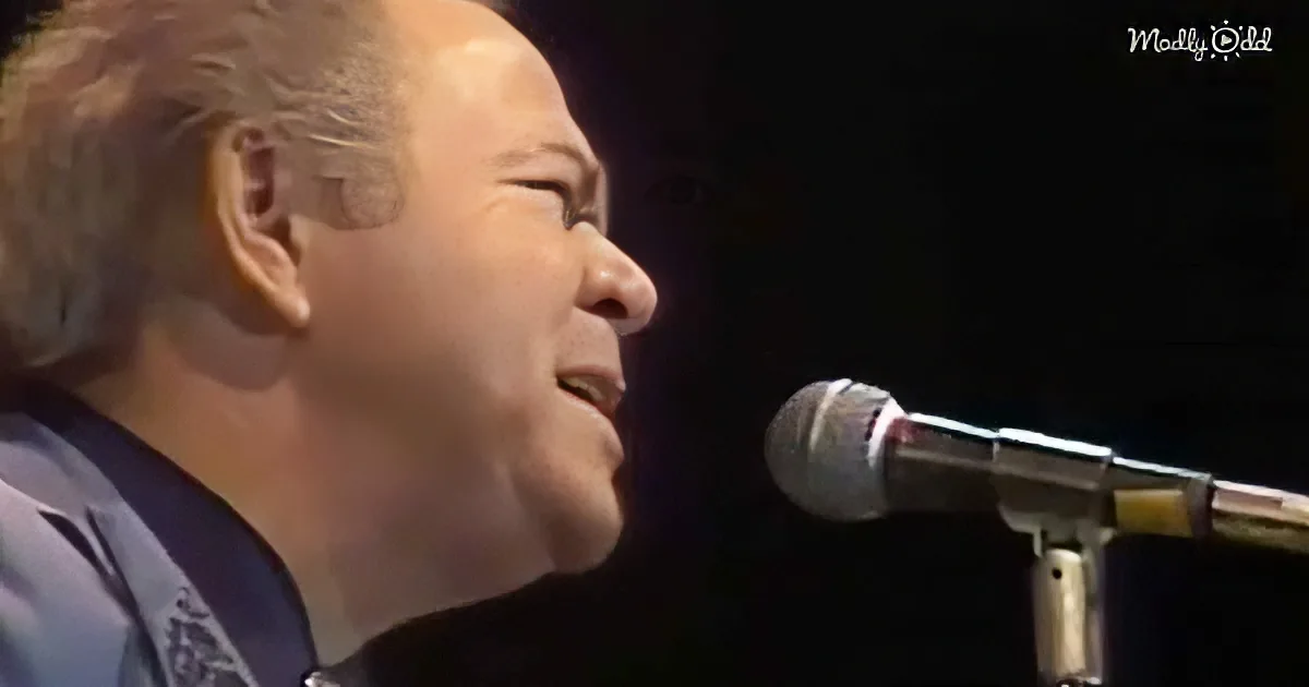 A man with short, light-colored hair sings into a microphone, his face partially illuminated.