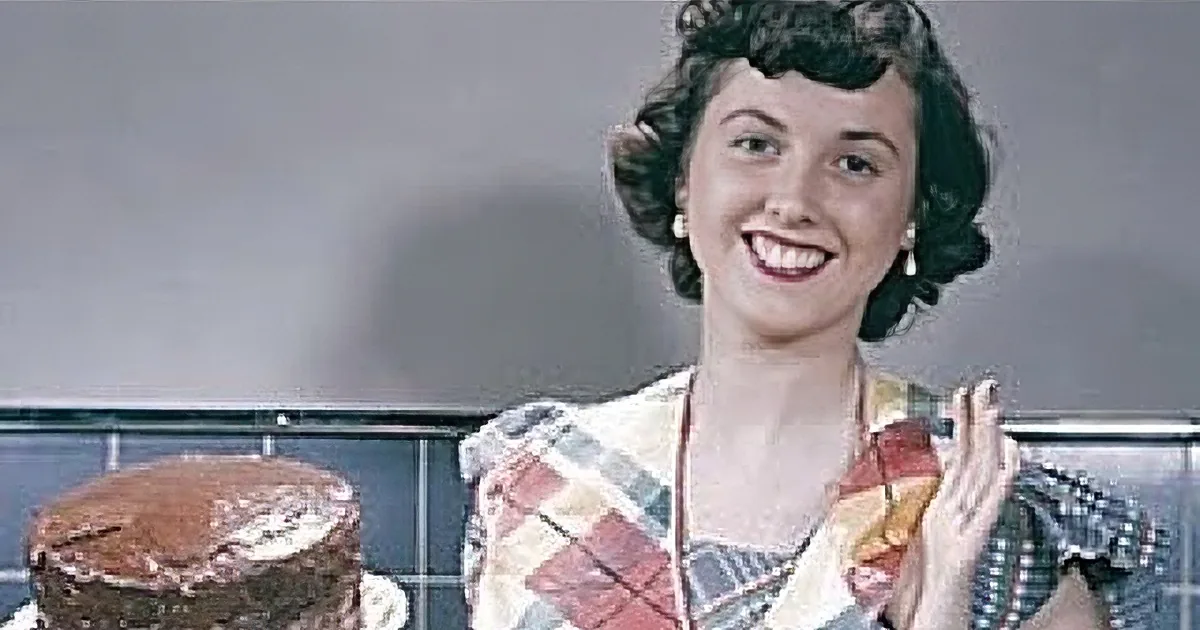 A woman with curly hair, wearing a blue top, gazes downward near yellow flowers.