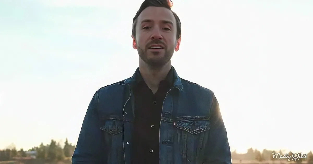 A man in a denim jacket and black shirt stands outdoors with a blurred background.