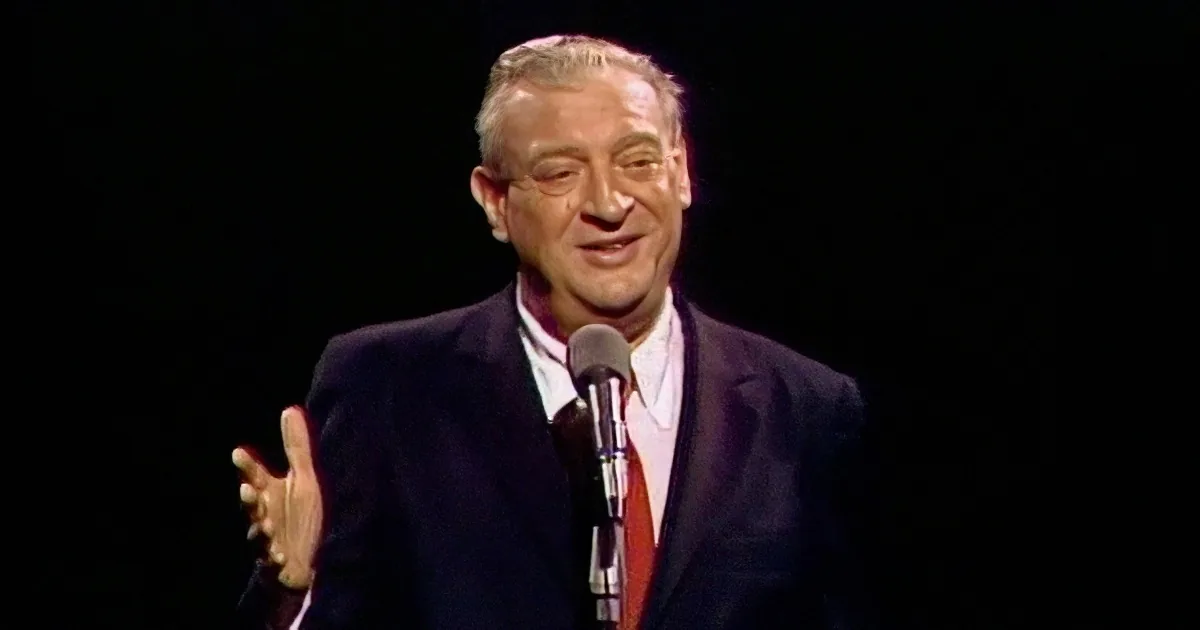 A man in a suit and tie speaks into a microphone, gesturing with his right hand.
