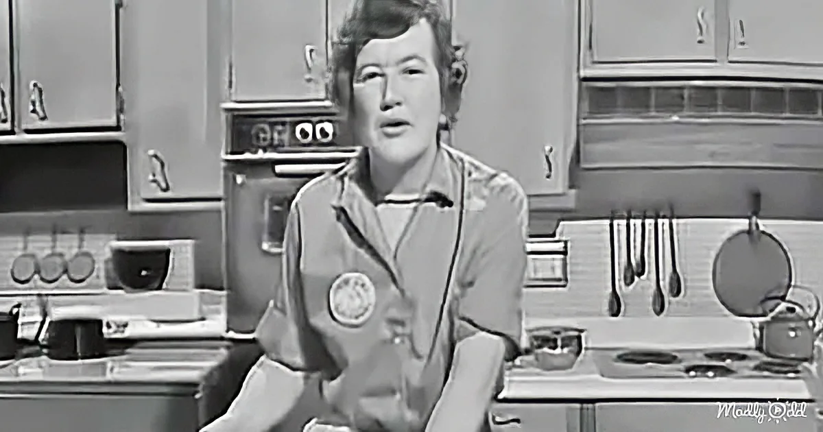 A woman in a kitchen, wearing a chef's jacket, sits with a stove and utensils in the background.