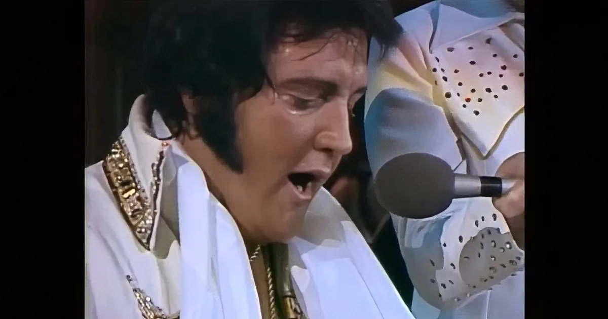 A performer in a white suit sings into a microphone, with "ELVIS" illuminated in red lights behind.