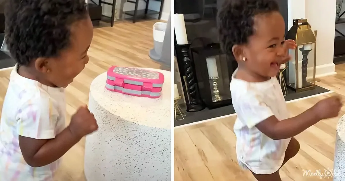 A toddler in a white tie-dye shirt is excitedly clapping and smiling at a pink and gray toy on a white speckled table.