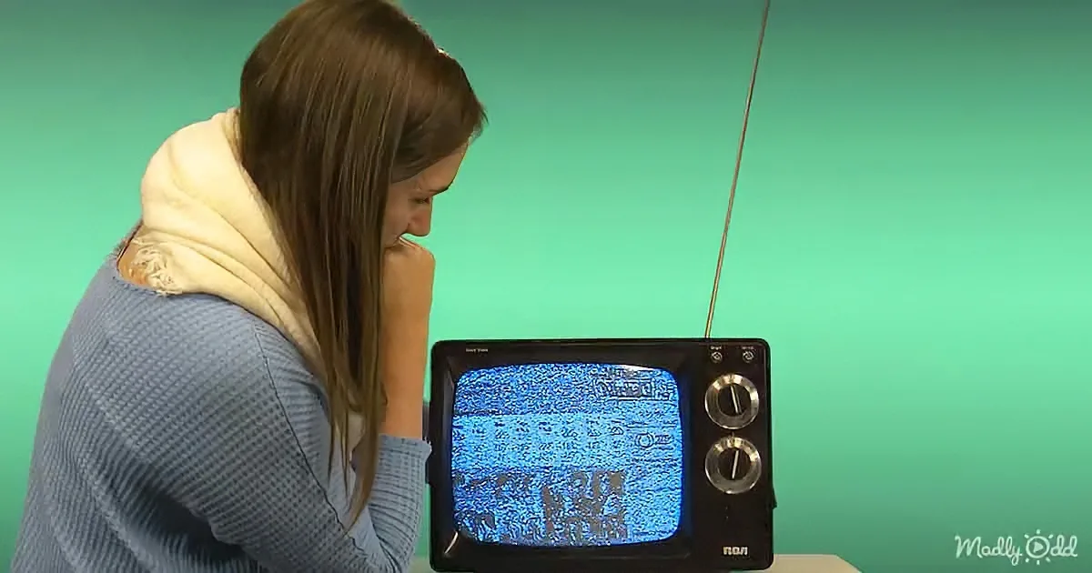 A woman in a blue sweater and white scarf leans on her hand, watching a vintage TV with static.