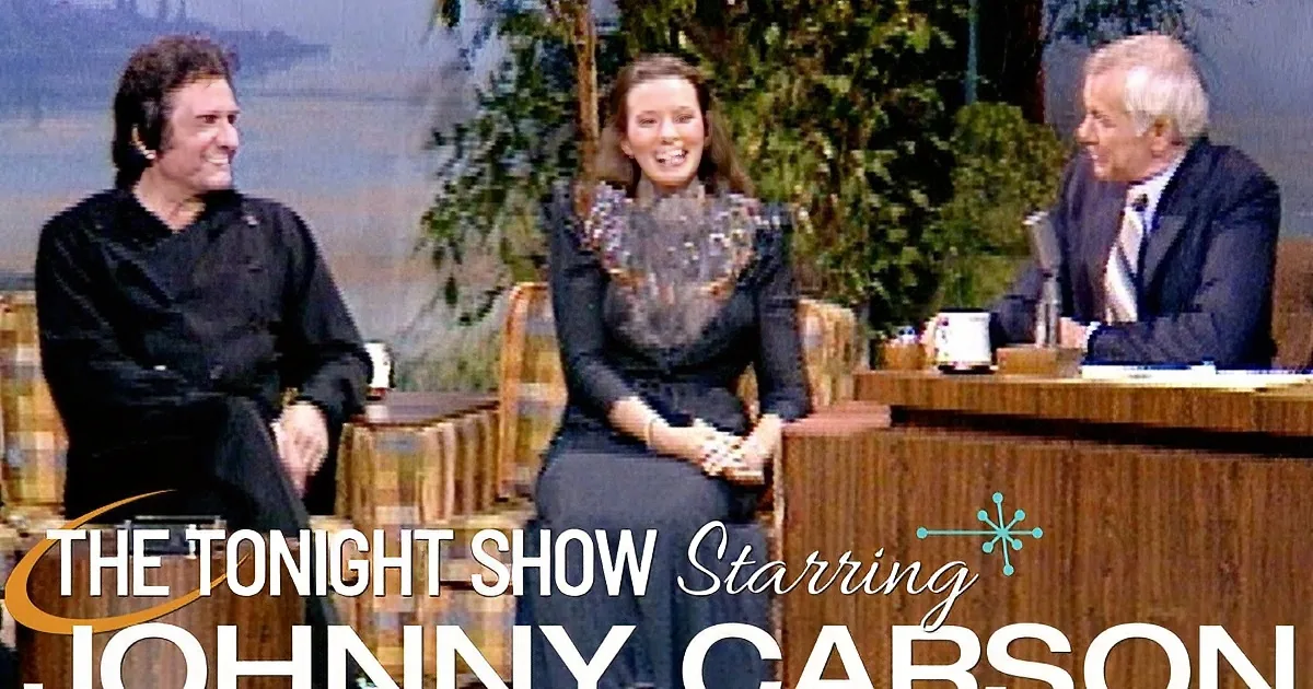 Johnny Cash in a black coat smiles while seated next to a man in a suit on a talk show set.