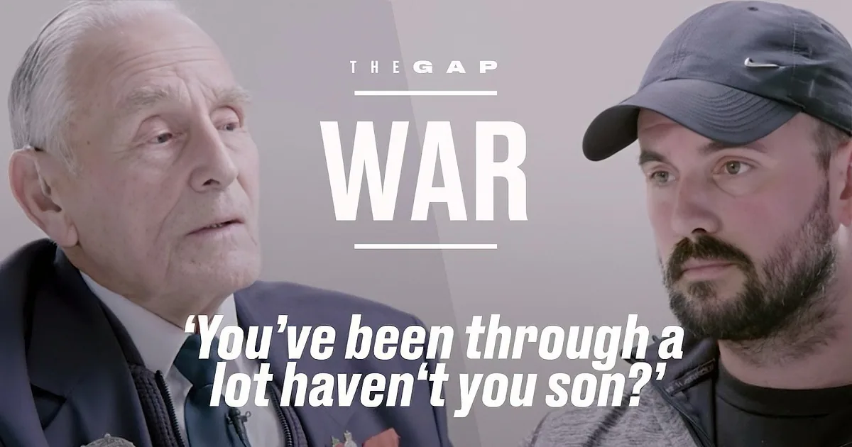 An older man in a suit and tie is speaking to a younger man wearing a cap and jacket.