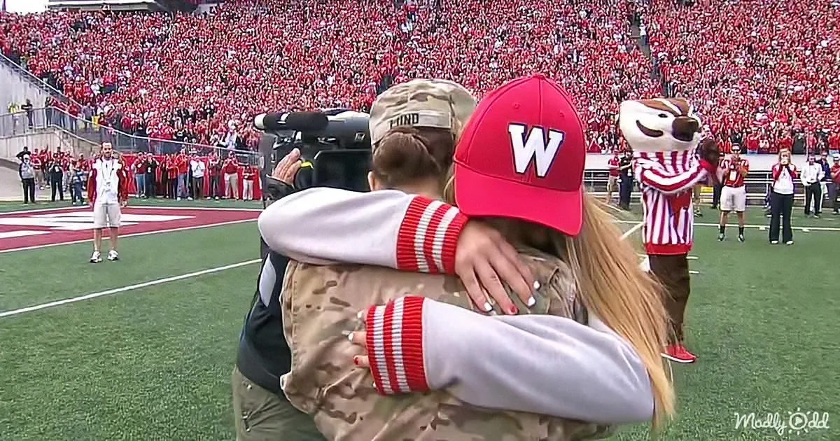 A soldier in uniform hugs a woman in a red "W" hat on a football field with a mascot and crowd in the background.