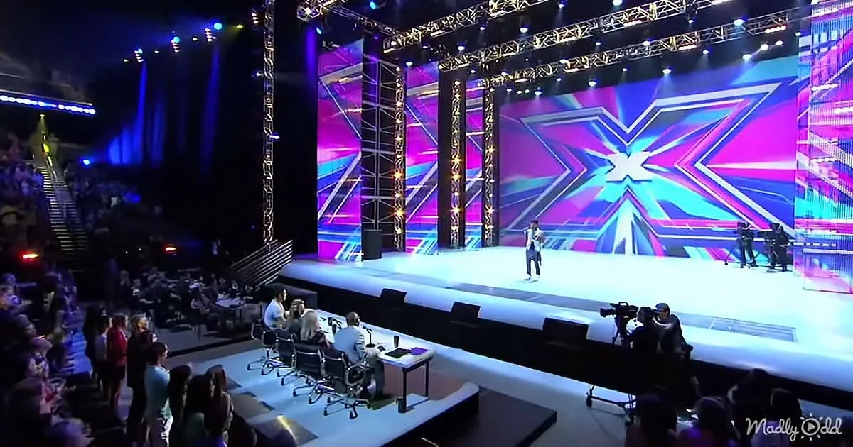 A performer stands on a brightly lit stage with a large "X" backdrop, surrounded by judges and an audience.
