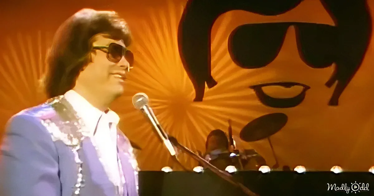 A man in a light blue suit and sunglasses smiles while playing a keyboard on stage.