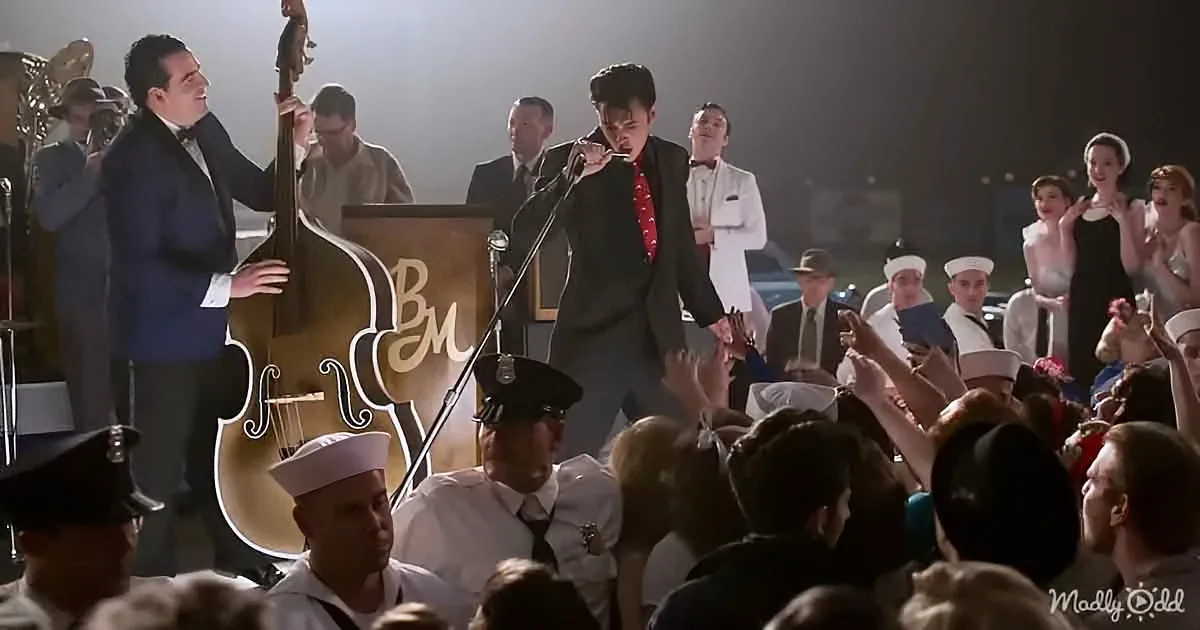 A man in a black suit sings into a microphone on stage with a band and audience.