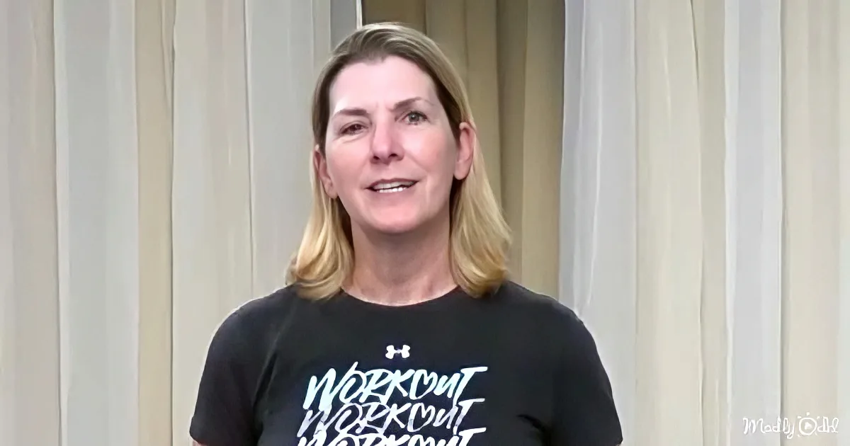 A woman with blonde hair, wearing a black Under Armour "Workout" t-shirt, stands in front of vertical blinds.