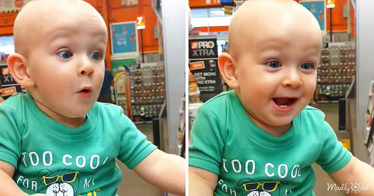 A baby in a green "Too Cool For School" shirt is smiling widely in a store setting.