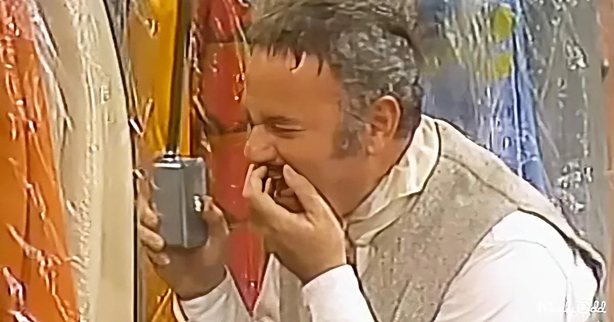 A man in traditional attire is holding a phone, appearing to be in distress or laughter, with his hand near his mouth.