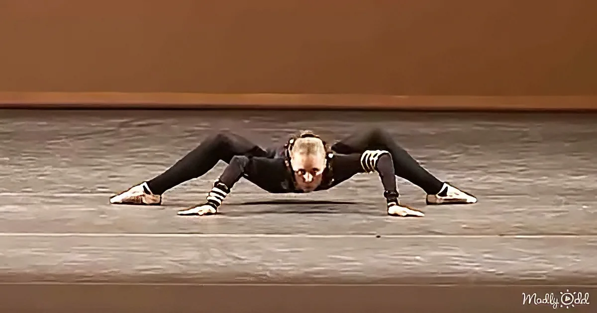 A person in a black outfit is performing a low, spread-out pose on a stage floor.