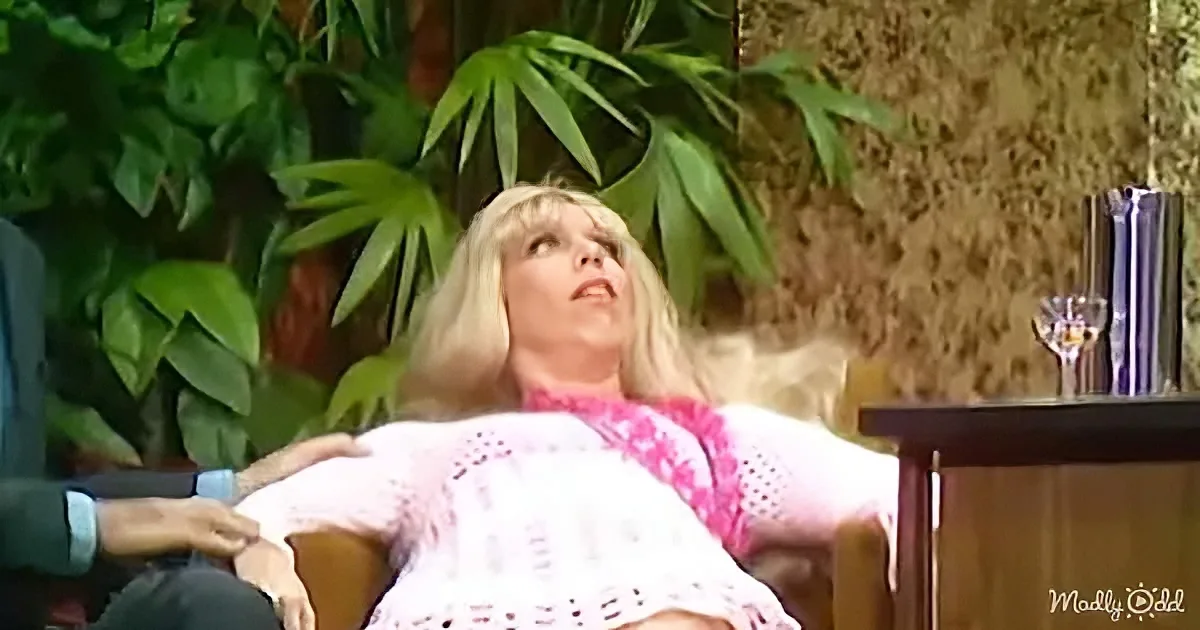 A woman with blonde hair, wearing a pink dress, reclines on a chair, looking up, with green plants and a textured wall in...