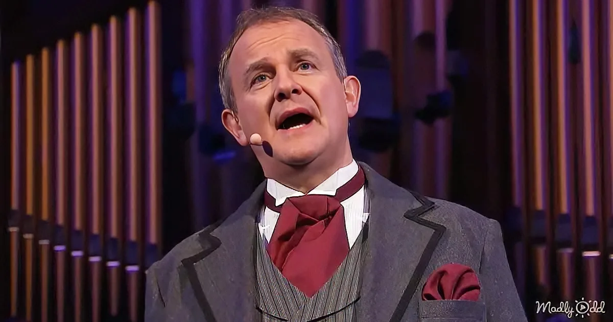 A man in a suit and red tie sings on stage with a microphone, against a backdrop of vertical pipes.
