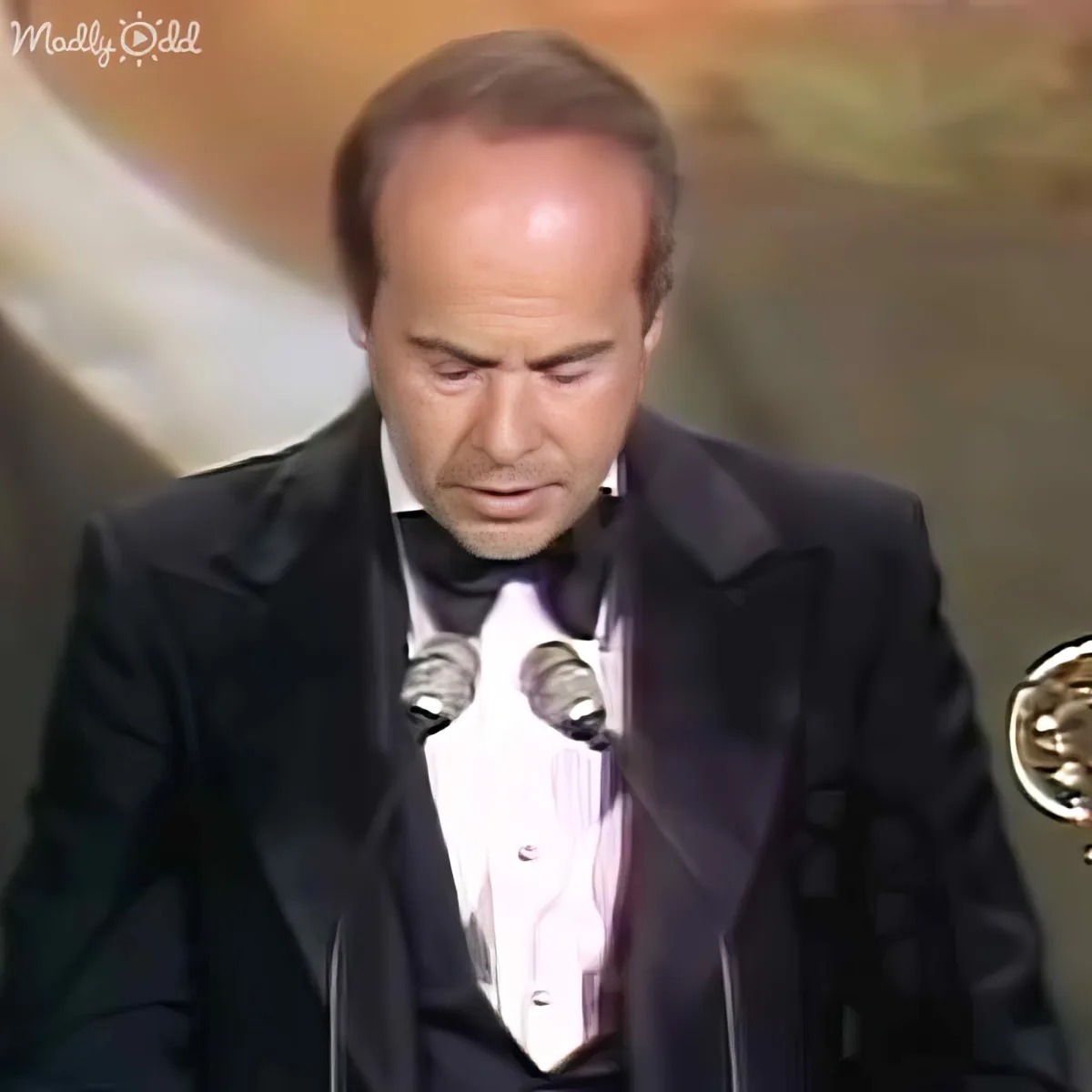 A man in a tuxedo stands at a podium, holding an award, looking down with a serious expression.