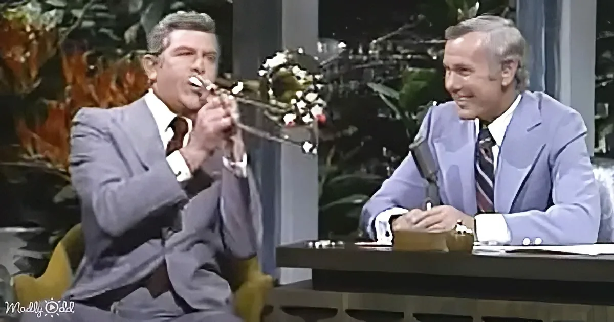 A man in a suit plays trumpet while another in a suit sits at a desk, both smiling.