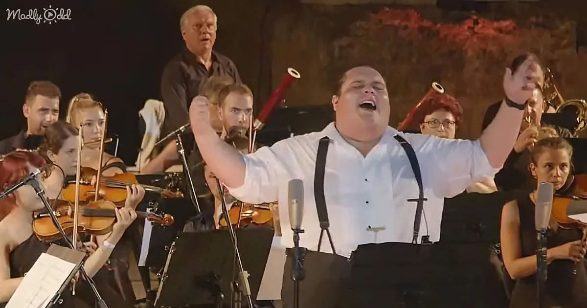 A man in white shirt and suspenders sings passionately in front of an orchestra.