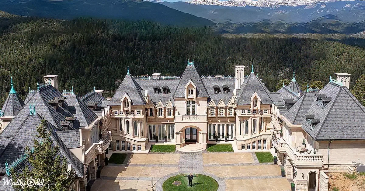 A grand, castle-like mansion with multiple towers and a courtyard, set against a backdrop of lush green hills and...