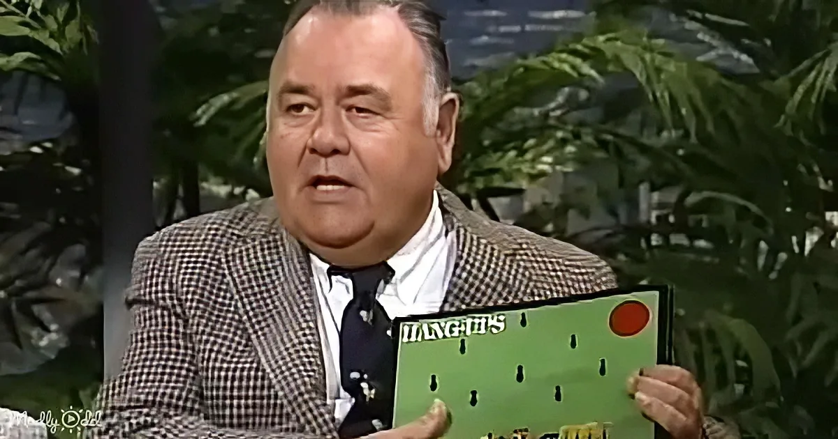 A man in a checkered suit holds a football playbook, surrounded by greenery.