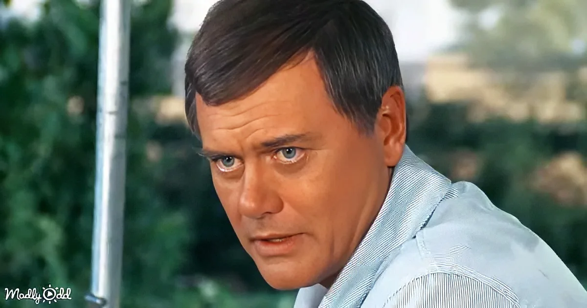 A man with short dark hair and a light blue shirt looks off to the side, with a blurred outdoor background.