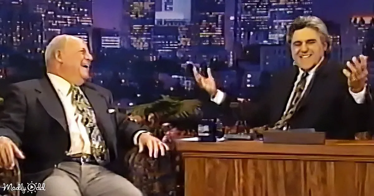 Two men in suits, one seated, the other behind a desk, both smiling and clapping, against a cityscape backdrop.