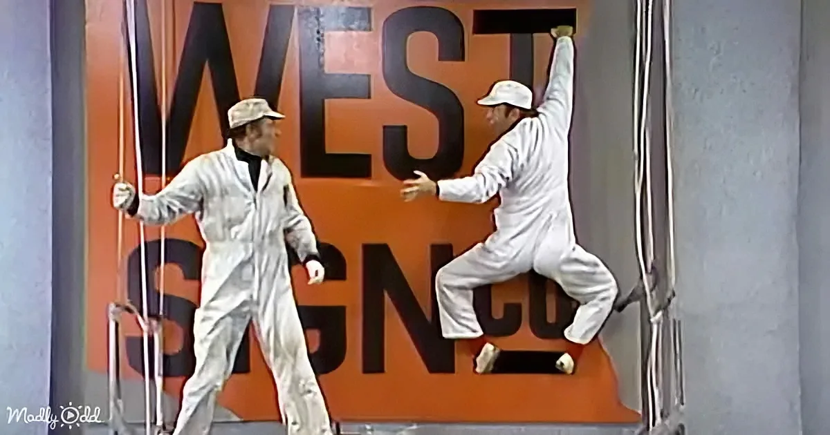 Two men in white coveralls are painting a large orange sign with the word "WEST" visible.