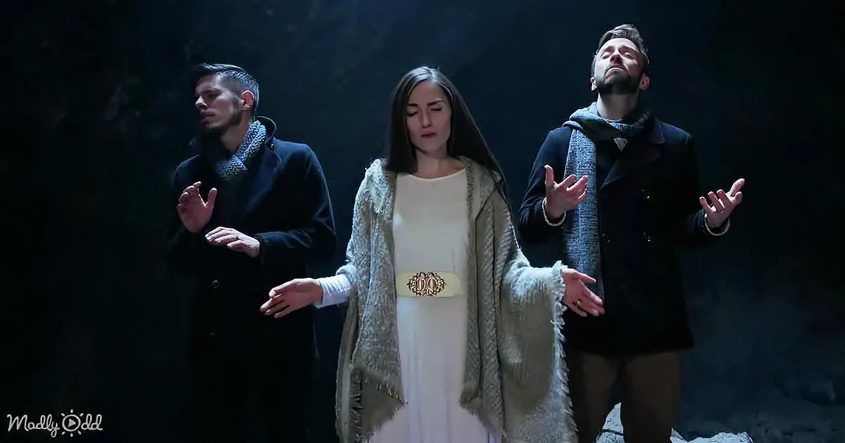 A woman in a white dress and beige shawl stands with her hands outstretched, flanked by two men in dark coats, all in a...