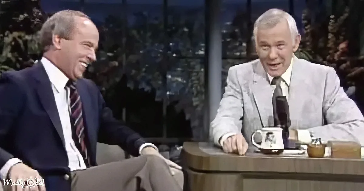 A man in a suit laughs while seated on a talk show set with another man in a light suit.