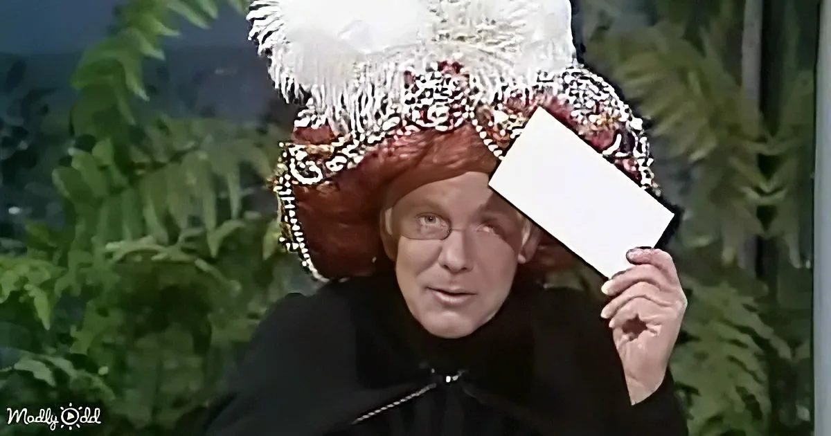 A person in a large, ornate hat holds a white card, with green foliage in the background.