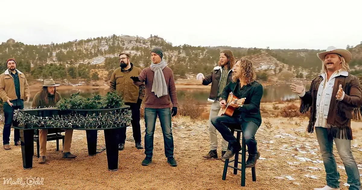 A group of people, including a guitarist, sing and play in a snowy, mountainous outdoor setting.