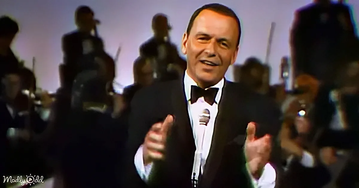 A man in a tuxedo sings into a microphone, gesturing with his hands, with a band in the background.