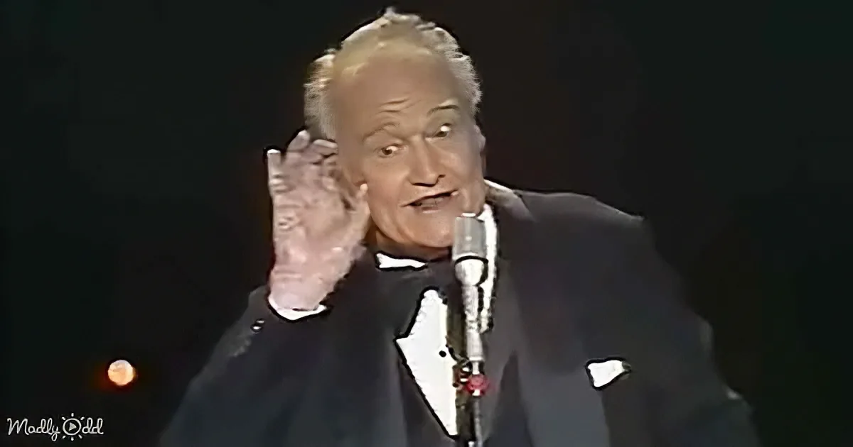 A man in a tuxedo gestures dramatically while speaking into a microphone on stage.